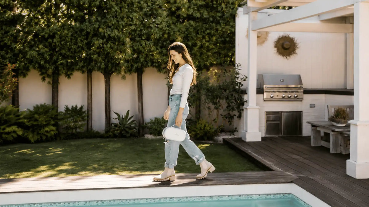 Woman walking by a backyard pool carrying a compact portable breast pump.