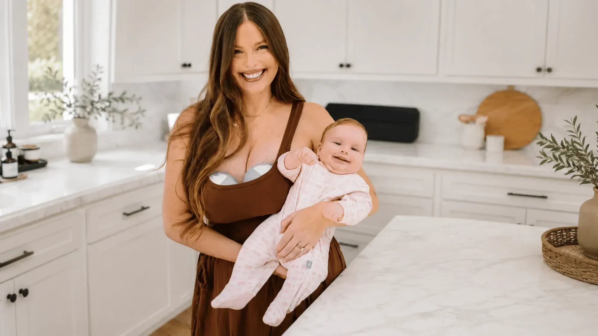 Smiling mother holding her baby in a bright kitchen while wearing a hands-free breast pump.