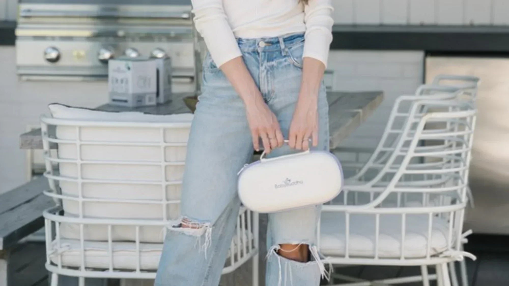 A woman in jeans holds a white BabyBuddha carrying case while standing beside an outdoor table and chairs.